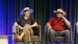Two people sitting on stage with a blue curtain background