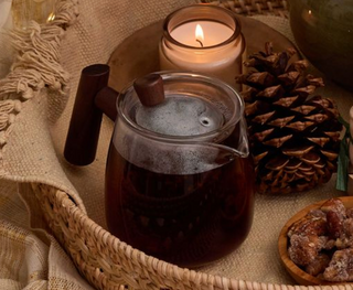 Glass teapot with steaming tea, lit candle, pine cones, and cookies on a textured surface.