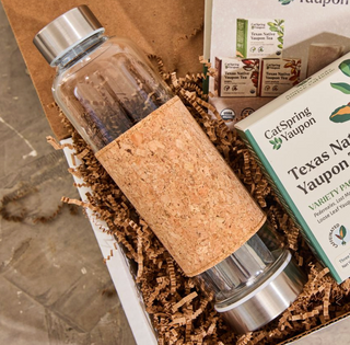 Glass water bottle with cork sleeve and metal cap in a gift box with Texas Native Yaupon tea packaging.